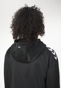 Persona con cabello rizado y negro de espaldas, vistiendo una sudadera con capucha negra con rayas de chevrones blancas en el hombro y pequeño logo blanco en la capucha.