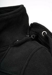Veste en polaire noire avec un col haut, dotée d'une poche zippée et d'accents en métal. Texture douce et coutures plates visibles.