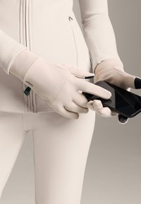 Jasnobeżowe rękawiczki z dotykowymi końcówkami palców, noszone przez osobę trzymającą smartfona. Gładka faktura, dopasowany krój.