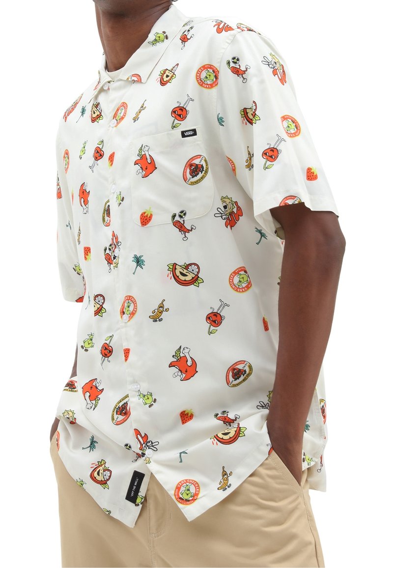 Chemise blanche à manches courtes boutonnée avec des imprimés colorés de fruits et légumes en dessin animé, portée par une personne ayant les mains dans les poches d'un pantalon beige.