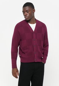Cardigan lavorato a maglia borgogna con motivo a quadri, scollo a V, chiusura con bottoni e due tasche frontali. Indossato sopra una camicia bianca.