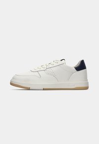Low-top white leather sneaker with beige sole, white laces, perforated side details, and navy heel accent.
