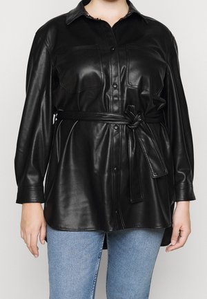 Kobieta nosząca czarną, zapinaną na guziki koszulę z eko-skóry z związanym paskiem w talii oraz niebieskie dżinsy, pokazana od szyi do bioder.