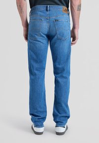 Mann trägt blaue Lee-Jeans und weiße Sneakers, steht mit dem Rücken zur Kamera vor einem schlichten hellen Hintergrund.