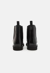Botine negre din piele cu un finisaj lucios, prezentând panouri laterale elastice, uși de tragere și tălpi plate din cauciuc. Viziune din spate arătată.