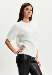 White short-sleeve cardigan made of fluffy knit material, featuring a round neck and several blue buttons, paired with black pants.