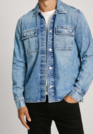 Hombre con chaqueta vaquera azul claro con bolsillos de solapa con botones sobre una camisa blanca y pantalones negros, una mano en el bolsillo.