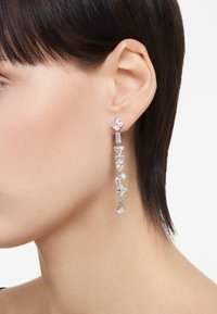 Långa silverörhängen med olika formade klara kristaller, inklusive fyrkantiga, triangulära och droppformade utformningar, satt i en linjär ordning.