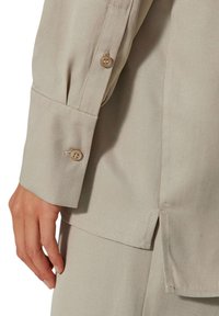Beige langärmliges Hemd mit Manschette und zwei Knöpfen, aus glattem Stoff mit seitlichem Schlitz für mehr Bewegungsfreiheit.