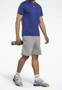 Modré sportovní tričko s krátkým rukávem a texturovanou látkou, doplněné šedými kraťasy. Drží modrou láhev na vodu a má na sobě vícebarevné sportovní boty.