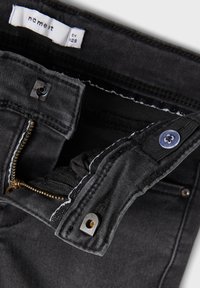 Pantalones de mezclilla negros con cierre de cremallera y botón. Presentan costuras en contraste y una etiqueta de tela en el interior de la cintura.