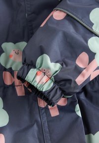 Navy rain jacket with elastic cuffs, featuring a playful pattern of smiling green clouds and pink accents. Made from a smooth, water-resistant material.