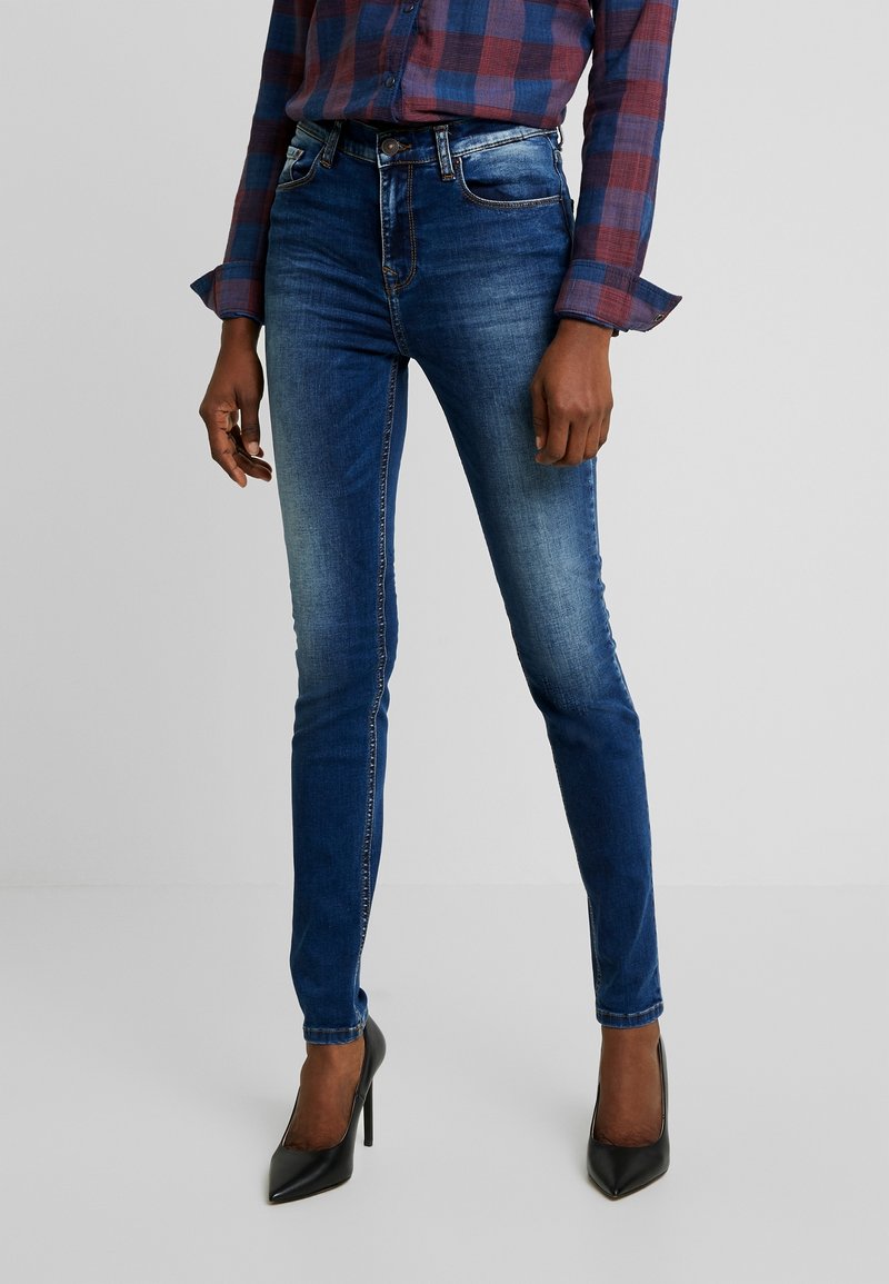 Jeans ajustados de denim azul oscuro con una textura suave, un ligero desgastado, trabillas para cinturón y bolsillos delanteros, combinados con zapatos de tacón negros.