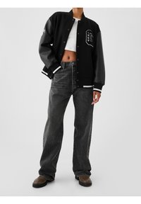 Veste universitaire noire avec manches en cuir, bandes blanches aux poignets et détail de lettre sur le devant ; associée à un jean noir taille haute et des chaussures marron.