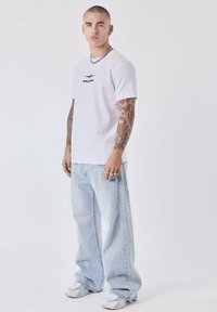 T-shirt bianca a maniche corte con logo nero sul petto, abbinata a jeans larghi blu chiaro e sneakers grigie, caratterizzata da anelli d'argento e una catena.
