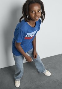 Dítě s dredy, oblečené v modrém tričku Levi's, světlých džínách a bílých teniskách, stojí proti světlému šedému zdi a tmavému šedému podlaze.