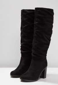 Botas altas hasta la rodilla de color negro y con caída, hechas de suave ante. Presentan un tacón grueso y una punta redondeada, con un diseño texturizado y fruncido a lo largo del cañón.