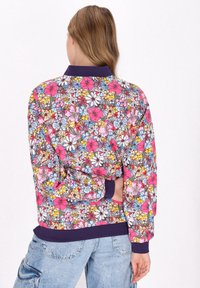 Bomberjack met bloemenprint, featuring roze, blauwe en gele bloemen op een donkere achtergrond. Marineblauwe ribkraag en -manchetten complementeren het ontwerp.