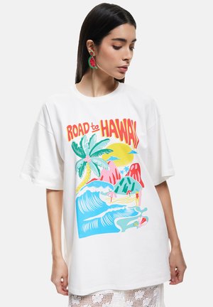 Mujer joven con pelo largo y oscuro, vistiendo una camiseta blanca oversize con un colorido gráfico playero de "Camino a Hawái" y pendientes en forma de sandía.