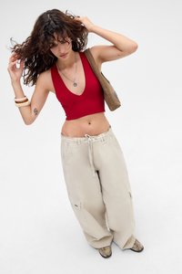 Žena u crvenom bezrukavom crop topu, bež širokim hlačama, smeškim cipelama sa zmijskim uzorkom i svetlosmeškom torbom na ramenu, prilagođava svoju kovrčavu kosu.