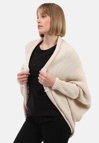 Jeune femme portant un haut noir et un pantalon noir, tenant un cardigan tricoté beige drapé sur ses épaules, regardant sur le côté.