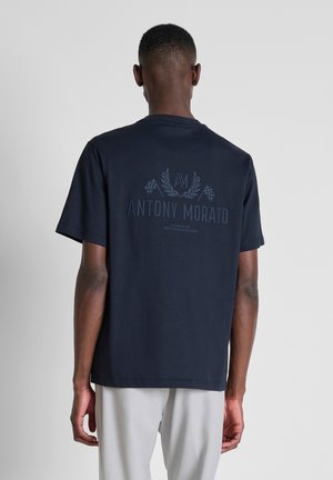 Man draagt een navyblauw Antony Morato t-shirt met logo en vlaggen op de achterkant, gecombineerd met lichtgrijze broek, staand tegen een effen achtergrond.