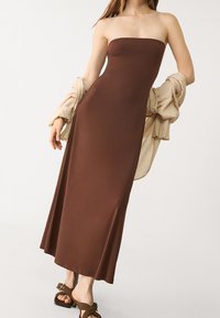 Bezramienna brązowa sukienka maxi z gładkiego materiału, dopasowana góra, zwiewna spódnica. Połączona z jasnobeżowym, oversize'owym topem i brązowymi sandałami.