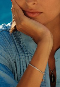 Srebrna zapestnica z zapletenim dizajnom, leži na zagoreli zapestju, na mehkemu modrem ozadju.