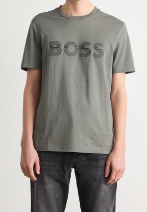 Persona que lleva una camiseta gris de manga corta con la palabra BOSS en grandes letras negras en el pecho y vaqueros gris oscuro.