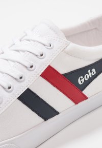 Sneaker in tela bianca con strisce rosse e navy. Presenta lacci bianchi, stampa del logo e una suola in gomma. Design semplice e a motivi.