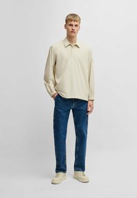 Helle beige Langarm-Polo mit einem geknöpften Kragen, kombiniert mit dunkelblauen Jeans und beigen Sneakers. Das Polo hat geraffte Ärmelabschlüsse.