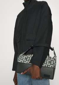 Borsa grigia e nera con un motivo geometrico, dotata di una base in pelle e tracolla regolabile. La borsa è tenuta in mano con una giacca testurizzata visibile.