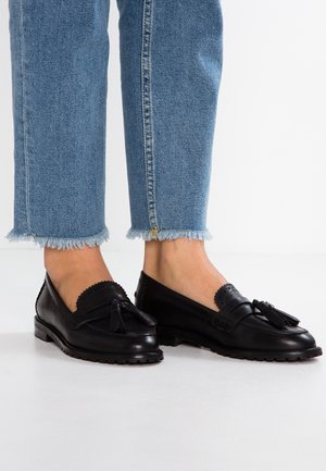 Persoon in blauwe jeans met gerafelde zoom en zwarte leren loafers met kwastjes, staande op een witte vloer.