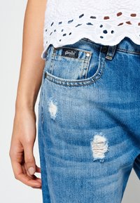 Roztrhané modré džíny z denimu s vybledlým umytím, s kapsou na nášivku a značkovou etiketou na pase. Textura je měkká a opotřebovaná.