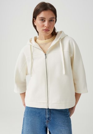 Frau mit kurzen braunen Haaren, die einen weißen Kapuzenpullover mit Reißverschluss und Kordelzügen, einen beigen Stricktop und blaue Jeans vor einfarbigem Hintergrund trägt.