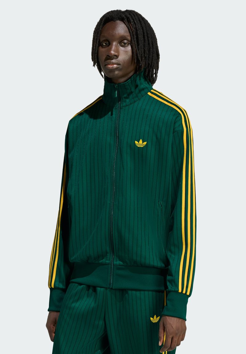 Zelená šušťáková bunda na zips s vertikálnymi pruhmi, žltými akcentmi a malým žltým logom. Má vysoký golier a elastické manžety.