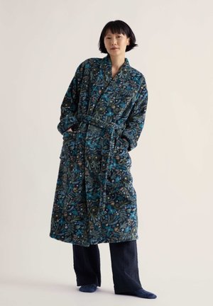 Personne portant une robe à motif floral bleu et vert avec une ceinture, debout les mains dans les poches devant un fond clair uni.