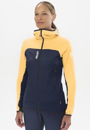 Femme portant une veste zippée jaune et bleu marine avec un col montant, debout devant un fond blanc uni.