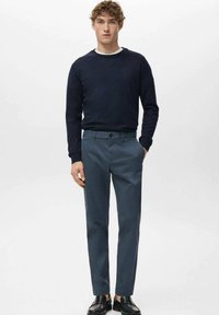 Young man stands with hands in pockets wearing navy blue sweater, slate blue trousers, and black loafers against white background.