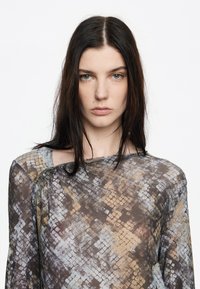 Jonge vrouw met lang donker haar, gekleed in een doorschijnende slangenprint top, die recht vooruit kijkt met een neutrale expressie op een witte achtergrond.