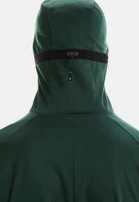 Dunkelgrüne Kapuzenjacke mit schwarzem verstellbarem Zugband über dem Rücken der Kapuze vor einem einfarbigen Hintergrund.