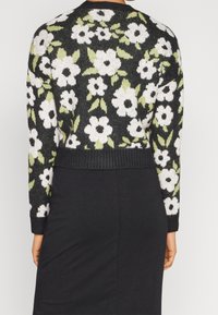 Strickjacke mit schwarzem Grundton und floralem Muster in Weiß und Grün, cropped Länge und gerippten Bündchen und Saum.