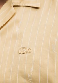 Chemise beige avec fines rayures verticales blanches, logo crocodile beige brodé sur la poitrine, et col pointu.