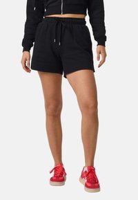 Frau trägt schwarze Shorts mit Kordelzug und rote Adidas-Sneaker, steht vor weißem Hintergrund.