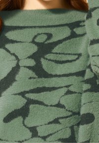 Zielony sweter o fakturze z miękką, puszystą powierzchnią i abstrakcyjnymi czarnymi wzorami. Wzór zawiera zaokrąglone kształty i różną grubość linii.