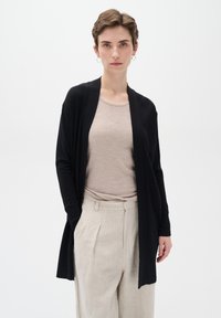 Zwarte cardigan met open voorkant en lange mouwen, gemaakt van een zachte stof. Gemaakt over een lichtbeige top en gecombineerd met lichte linnen broek.