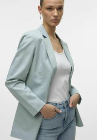 Blazer azzurro chiaro con vestibilità sartoriale, dotato di revers a lancia, texture liscia e maniche lunghe, indossato sopra una maglietta bianca e jeans.