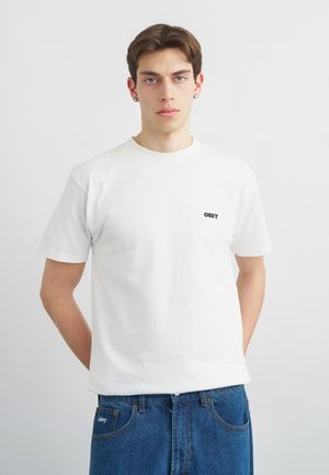 Νεαρός άνδρας με κοντά καστανά μαλλιά, που φοράει λευκό μπλουζάκι OBEY και μπλε τζιν, σε απλό γκρι φόντο.