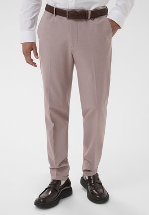 Mężczyzna ubrany w jasnobeżowe, dopasowane spodnie, ciemnobrązowy skórzany pasek, białą koszulę, białe skarpetki oraz ciemnobrązowe mokasyny, stojący z rękami w kieszeniach.