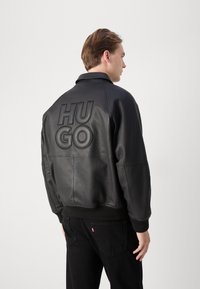 Melns ādas bumbierīša jaka ar izreljefu "HUGO" zīmolu aizmugurē, ribotām piedurknēm un apkakli. Valkāta kopā ar melnām biksēm.